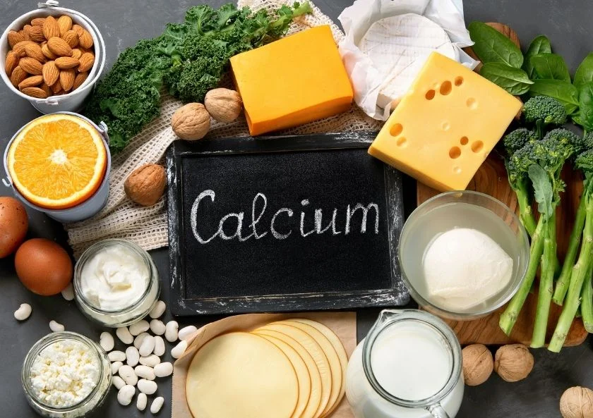 Calcium-rich foods including cheese, milk, nuts, eggs, leafy greens, and orange around a chalkboard labeled ‘Calcium.
